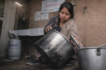 Mujer agachada de perfil, vertiendo de una olla metálica grande a otra. Viste ropa estampada y un chal. Detrás hay un cilindro de gas y un letrero que dice "Bienvenidos".