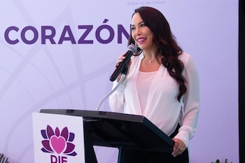 Una mujer con blusa blanca habla frente a un micrófono en un atril con el logotipo del DIF y un fondo con texto morado