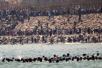 Numerosas personas en el mar y en la orilla rocosa. Al fondo, una valla con alambre de púas en lo alto de un muro de tierra