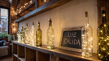 Botellas de vidrio con luces en su interior sobre un estante de madera junto a una pequeña pizarra con el texto luz cálida escrito con tiza.