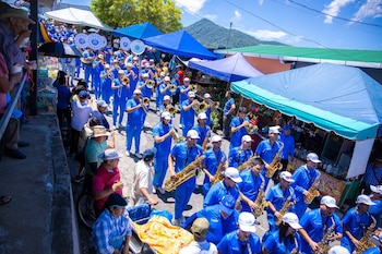 Hombre y mujer asiáticos sonriendo en primer plano, la mujer sostiene un letrero con forma de piña que dice "Feria de la Piña 2026 Santa María Ostuma"