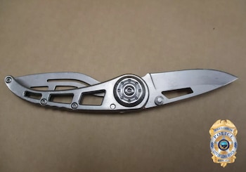 Cuchillo plegable metálico plateado con hoja extendida sobre fondo marrón claro. El escudo del Departamento de Policía de Long Beach se ve en la esquina inferior derecha