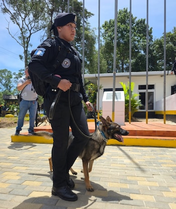 Agente de policía mujer con uniforme negro y boina negra, sujetando una correa, camina junto a un perro pastor alemán con collar azul en un exterior pavimentado
