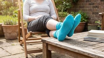 Piernas de una persona con calcetines azules brillantes descansando sobre una mesa de madera en un patio. Al fondo, un jardín con macetas y una pared de ladrillo.