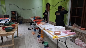 Dos agentes antinarcóticos inspeccionan una habitación con mesas llenas de recipientes y paquetes de droga blanca y rosa, con microondas apilados al fondo