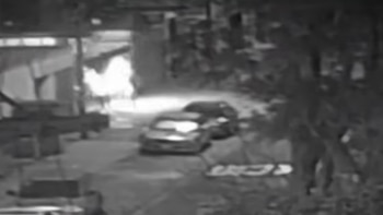 Fotografía en blanco y negro de una calle oscura con vehículos estacionados y un resplandor de fuego que ilumina una zona distante