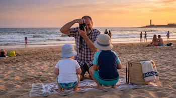 Hombre sonriente con cámara, dos niños de espaldas con sombreros en la arena, toalla, cesta, mar, faro y personas al fondo durante atardecer.