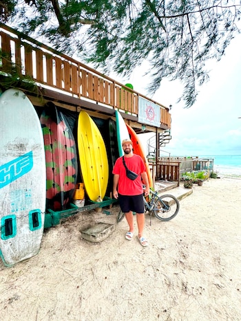 Hombre sonriente con camiseta roja, bermudas y gorra de pie en la arena. Detrás, tablas de surf, bicicleta, edificio de madera y mar turquesa