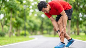 Un hombre corredor vestido con camiseta roja y pantalones cortos negros se agarra la tibia con dolor en una pierna, en un camino dentro de un parque.