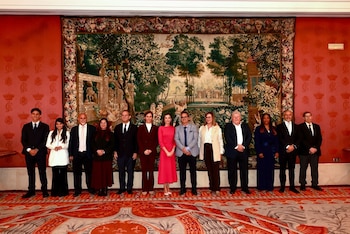 Trece personas, incluyendo a la Reina Letizia en el centro, posan de pie frente a un gran tapiz de paisaje y una pared roja decorada con motivos de coronas doradas