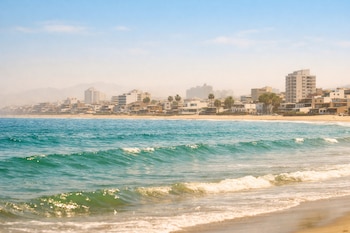 Playa peruana con olas azul verdoso en primer plano. Al fondo, una ciudad costera con edificios y palmeras, ligeramente difuminada por el brillo del intenso sol.