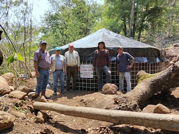 Cinco hombres posan frente a una estructura cilíndrica con malla y lona. El terreno es rocoso con árboles frondosos y troncos caídos