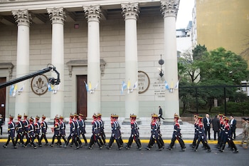 Soldados con uniformes de gala oscuros y sombreros rojos marchan por una calle. Al fondo, un edificio clásico con columnas, adornado con banderas y una grúa de cámara