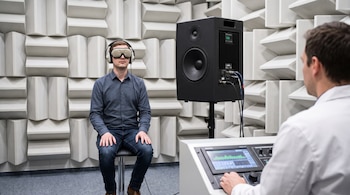 Un hombre usa gafas opacas y auriculares en una habitación con paneles acústicos, junto a un altavoz negro y un técnico con una consola de control.