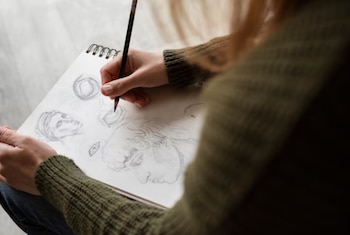 Manos de una persona sosteniendo un lápiz negro sobre un cuaderno de bocetos. El cuaderno muestra dibujos de rostros, ojos y formas geométricas