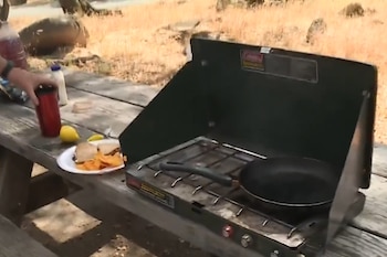 Una cocina portátil con una sartén sobre una mesa de madera de campo junto a un plato con un sándwich y un vaso rojo