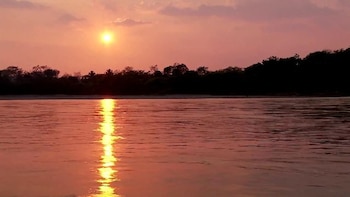 Sol redondo, cielo anaranjado, nubes rosadas, río con reflejo solar brillante, orilla con árboles silueteados