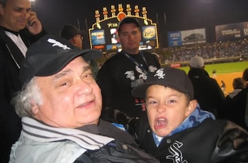 Primer plano de un hombre de cabello blanco sonriendo y un niño haciendo una mueca, ambos con gorras de los Chicago White Sox, en un estadio nocturno