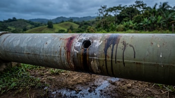 Primer plano de una tubería metálica industrial con un orificio de perforación del que gotea un líquido oscuro. Al fondo se ve un paisaje rural con vegetación.