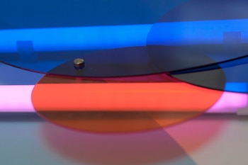 Luces de neón azul y rosa iluminan formas circulares transparentes de colores rojo y azul superpuestas, con reflejos y un pequeño tornillo dorado