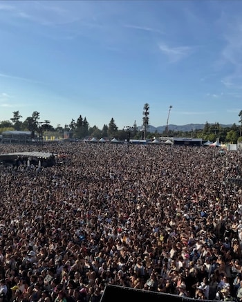 Vasta multitud de personas al aire libre, bajo un cielo azul con nubes claras. Hay árboles, tiendas de campaña y estructuras de metal en el fondo