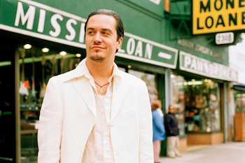 Hombre con cabello oscuro y barba, vestido con blazer blanco y camisa clara, en primer plano. Tienda con letreros "Mission Loan" y "Pawnbrokers" al fondo