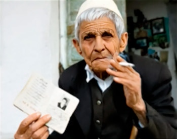 Persona mayor con gorro blanco y saco oscuro sostiene un documento de identidad abierto en una mano y un cigarrillo encendido en la otra