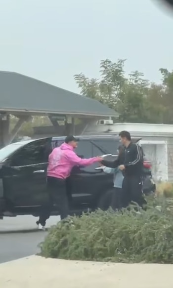 Dos hombres intercambiando algo cerca de un SUV negro. Uno lleva sudadera rosa, el otro chándal negro. De fondo, una casa y vegetación