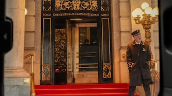 Un hombre con uniforme y gorra junto a una puerta oscura con adornos dorados, escalones con alfombra roja y una farola encendida