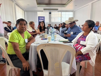 Varias personas, mesas, sillas, botellas de agua, tazas, cuadernos, bolígrafos, un cartel, ventanas con persianas verticales