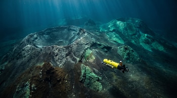 Un vehículo submarino autónomo amarillo y negro navega sobre un lecho marino con grandes calderas volcánicas bajo agua azul profunda con haces de luz.