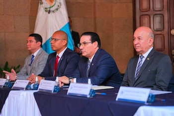 Cuatro hombres en trajes sentados en una mesa con manteles, rótulos de nombres, botellas de agua y gel antibacterial. La bandera de Guatemala está de fondo