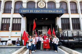 Un grupo de personas con banderas rojas protesta frente a la Corte de Constitucionalidad en Guatemala, un edificio blanco con arcos y ventanas oscuras