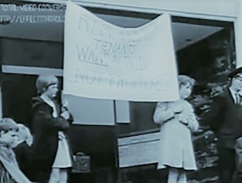 Grupo de personas, incluyendo niños, protesta al aire libre. Un cartel grande dice: 'TENANTS WANT ACTION NOT PROMISES'
