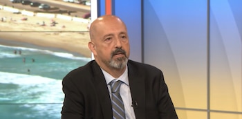 Hombre calvo con barba gris, traje oscuro, camisa celeste y corbata rayada. Al fondo, playa con personas y mar; estudio de televisión con paneles