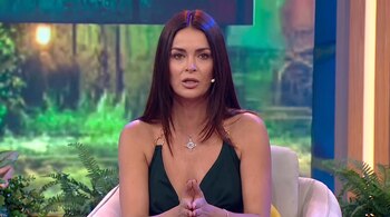 Una mujer de cabello oscuro con top verde y collar, sentada en un sillón claro, mira al frente con las manos juntas. De fondo, una pantalla con follaje borroso y plantas.