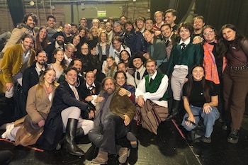 Un gran grupo de personas sonrientes, varias vestidas con atuendos de época, posan juntas en un escenario oscuro, algunas sentadas y otras de pie