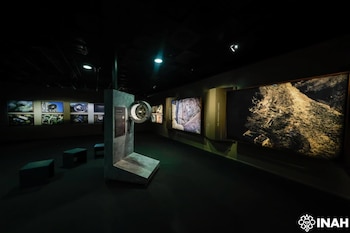 Sala oscura, seis fotografías en pared izquierda, dos fotografías grandes en pared derecha, módulo expositor con esfera central, tres bancos cuadrados