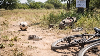 Zapatos de niño de 7 años, una pelota de fútbol, mochila y bicicleta en un camino de tierra. Un cartel de "SE BUSCA: AYUDA" está clavado en un árbol.