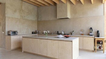Cocina moderna y espaciosa con gabinetes de madera clara, encimeras de concreto gris, paredes beige. Se ve una isla central con frutas, un florero, y electrodomésticos.