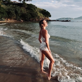 Mujer de espaldas con vestido blanco semitransparente, parada en la orilla del mar con el sol en la cara, y fondo de colinas verdes e islas