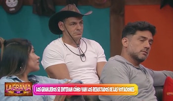 Mark Vito, con sombrero de vaquero y camiseta blanca, está sentado en un sofá junto a una mujer y otro hombre; su expresión es seria y contemplativa
