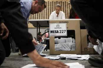 Primer plano de personas inclinadas, organizando papeletas electorales frente a una mesa con cajas de votación etiquetadas "MESA 227" y "SENADO"