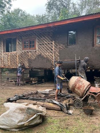 Tres personas construyen paredes de barro en una casa de madera. Hay una hormigonera, cubos, sacos de material y tierra. Fondo con árboles
