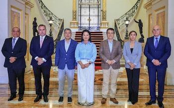 Siete personas, una mujer y seis hombres, de pie en un pasillo con escalinata al fondo, alfombra roja, estatuas y lámparas