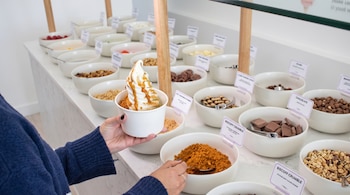 Persona sosteniendo un vaso de yogur helado con salsa y crumble. Un mostrador exhibe múltiples cuencos blancos con toppings variados y etiquetas de descripción.