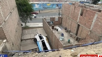 Una combi blanca y azul encallada entre muros de ladrillo sobre el techo de calamina de una casa, vista desde una pendiente de tierra