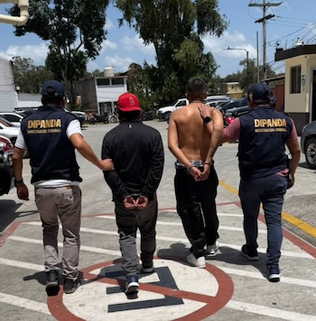 Dos agentes con chalecos de DIPANDA escoltan de espaldas a dos hombres esposados caminando en un estacionamiento