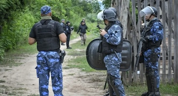 Grupo de policías con uniformes camuflados azules, chalecos tácticos, cascos y escudos en un camino de tierra junto a vegetación densa y una cerca de madera