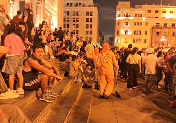 Vista nocturna de una calle concurrida en Lima; un trabajador de limpieza con uniforme naranja barre, mientras personas caminan y se sientan en escalinatas cerca de edificios iluminados